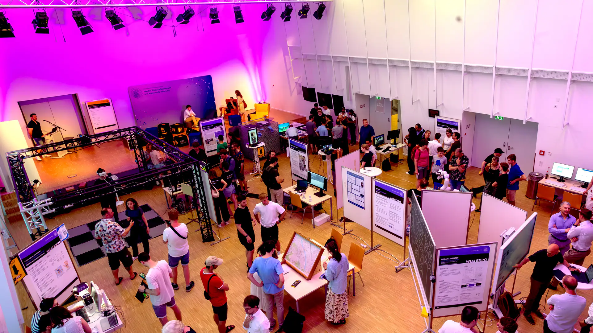 A packed exhibition hall seen from above: visitors walk between posters, screens and hands-on stations under violet stage lighting