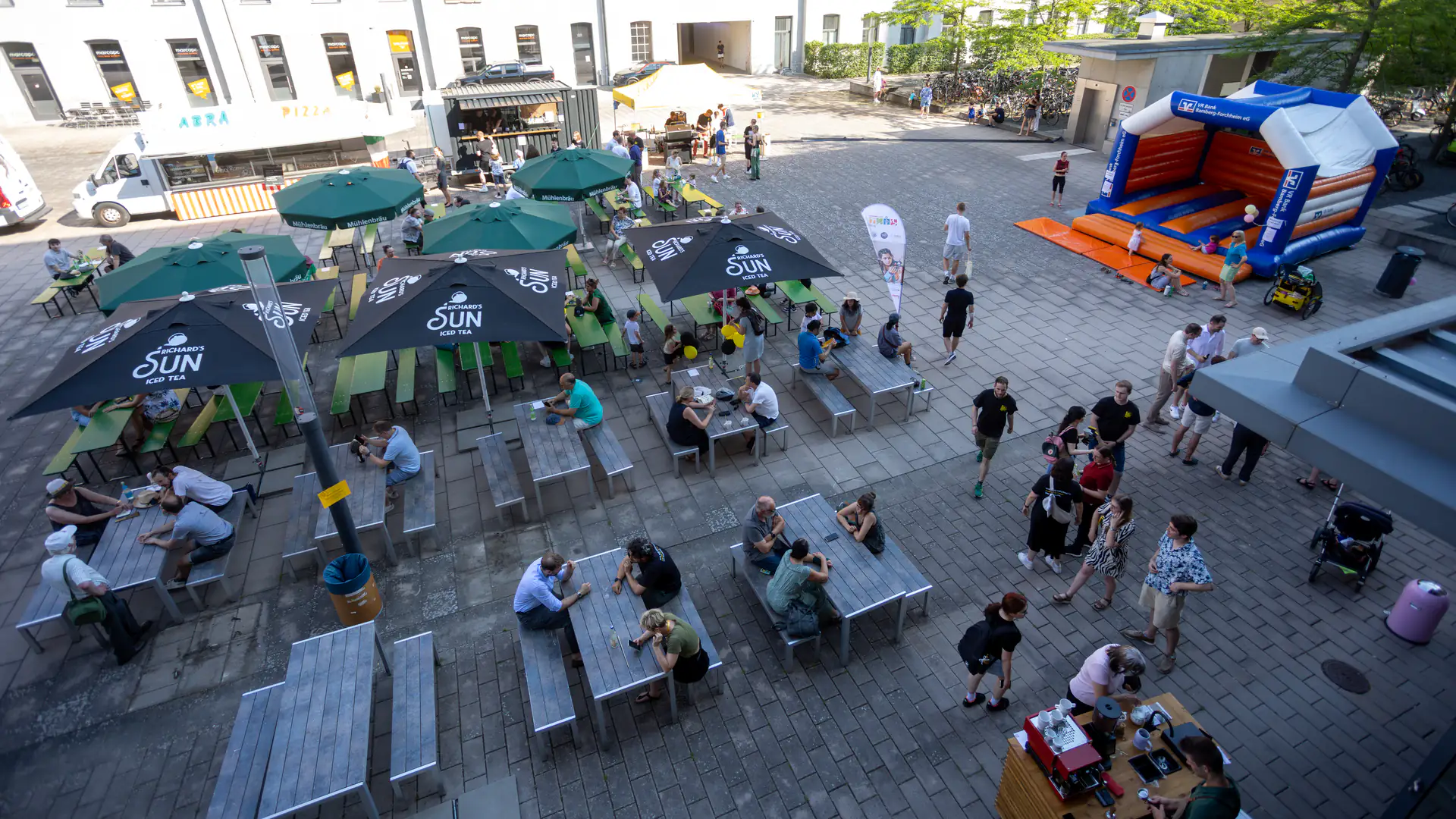 A sunny courtyard seen from above: people at long tables under parasols, a pizza van, a coffee stand and a bouncy castle