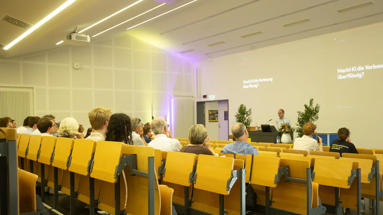 An audience of all ages in a bright lecture hall, seen from the back rows; a speaker stands at the front
